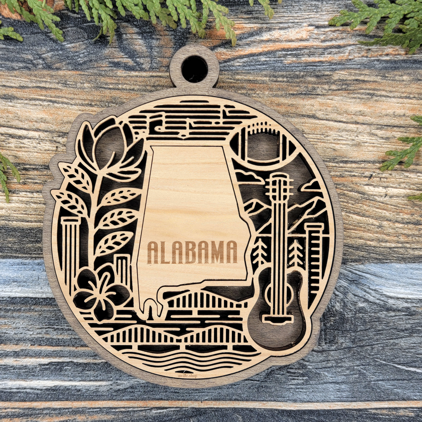 Layered Alabama wood ornament with a laser cut magnolia bloom, football, guitar and river bridge framing the engraved state shape