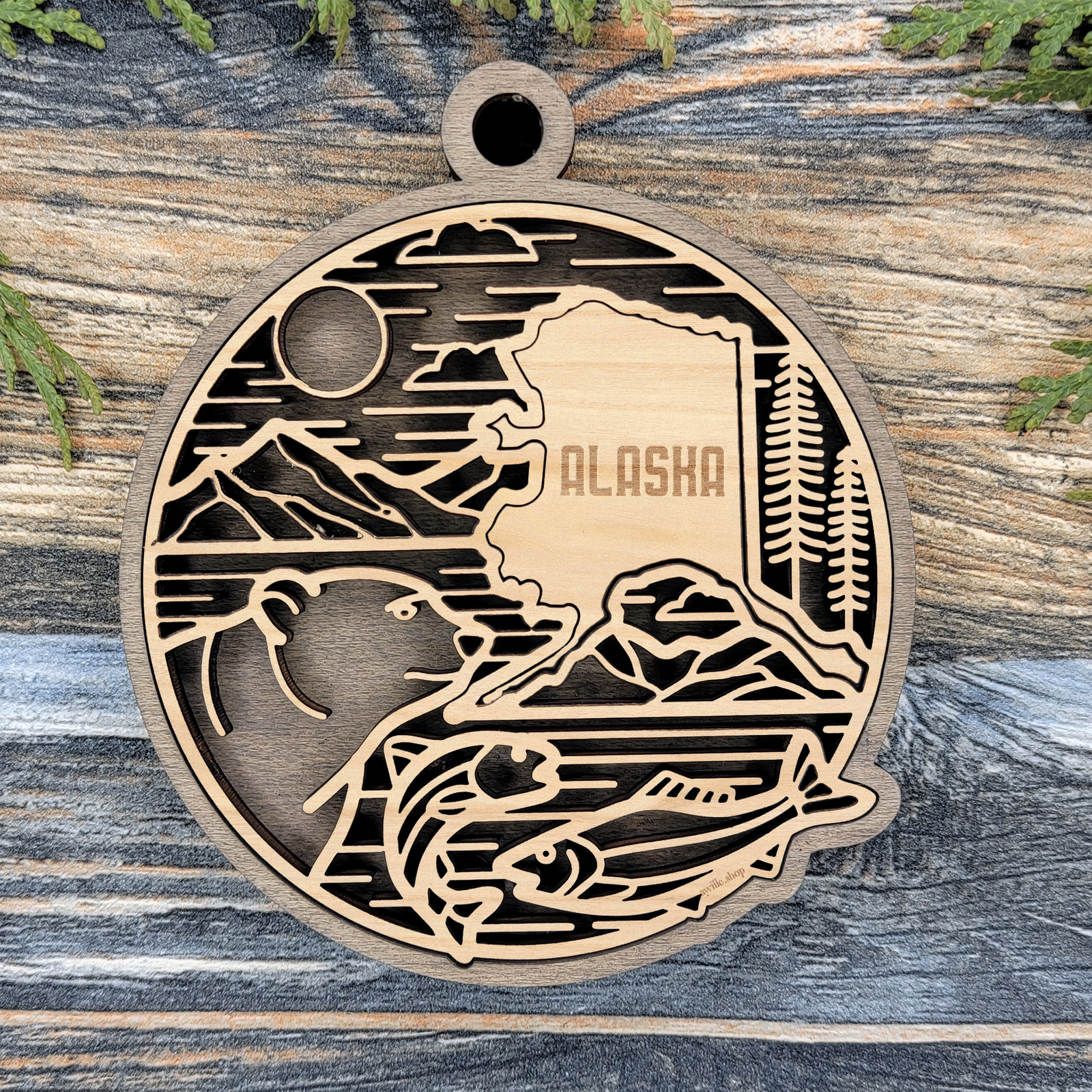 Layered Alaska wood ornament with a laser cut mountain range and evergreen trees framing the engraved state shape