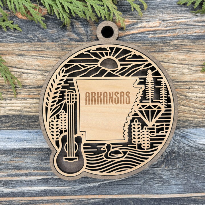 Layered Arkansas wood ornament with a laser cut guitar, pine trees, diamond and river framing the engraved state shape