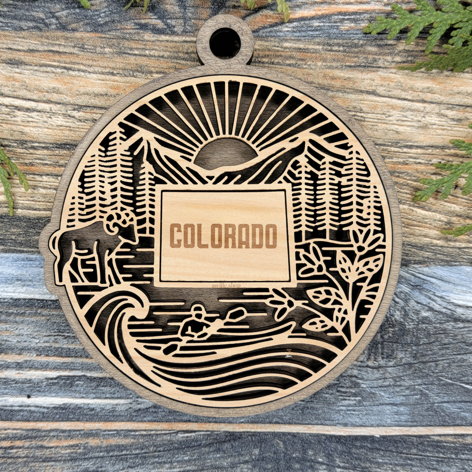 Layered Colorado wood ornament with a laser cut sunrise, mountains, pine trees, a bison and a kayaker framing the engraved state shape