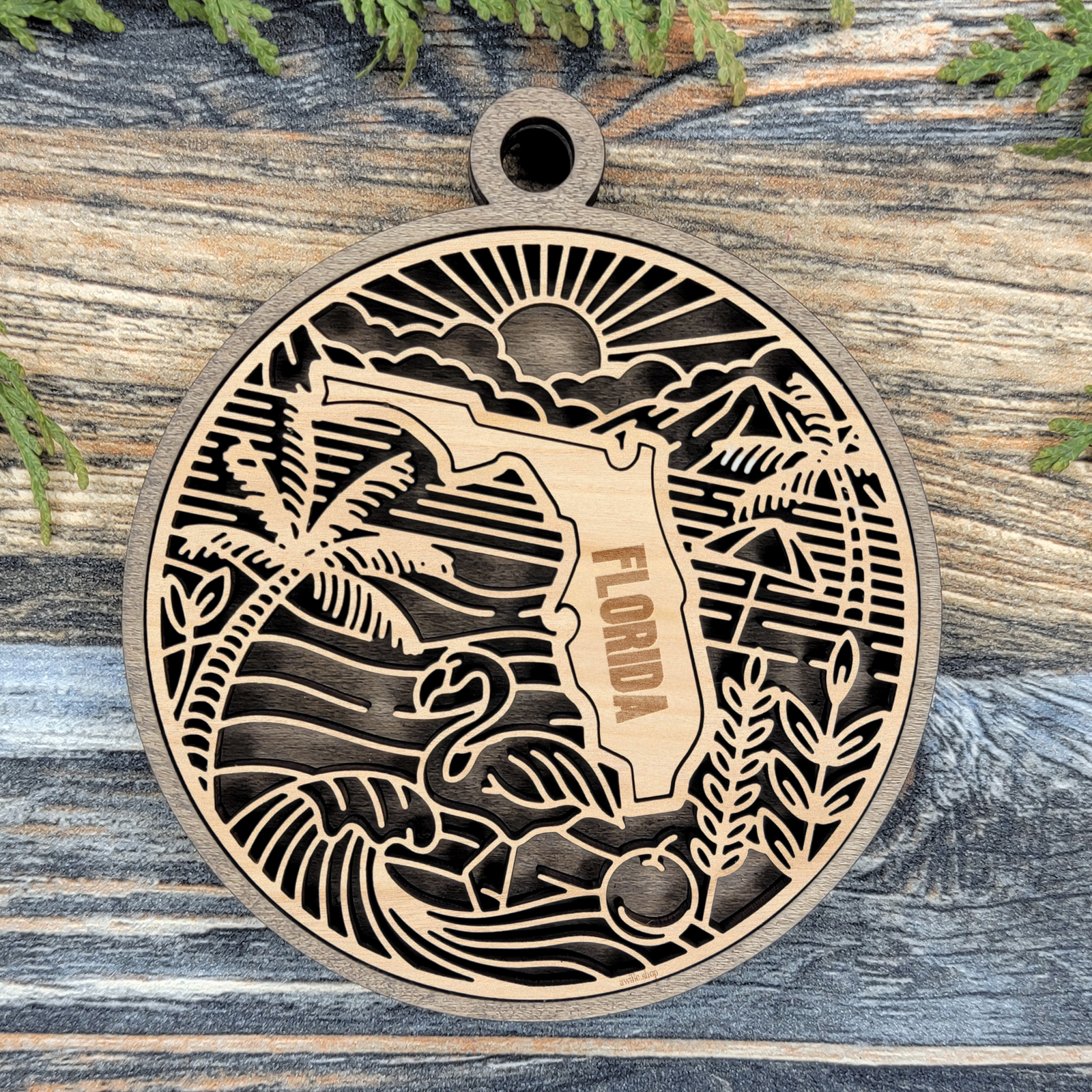 Layered Florida wood ornament with a laser cut sunrise, palm trees, a flamingo, an orange and ocean waves framing the engraved state shape