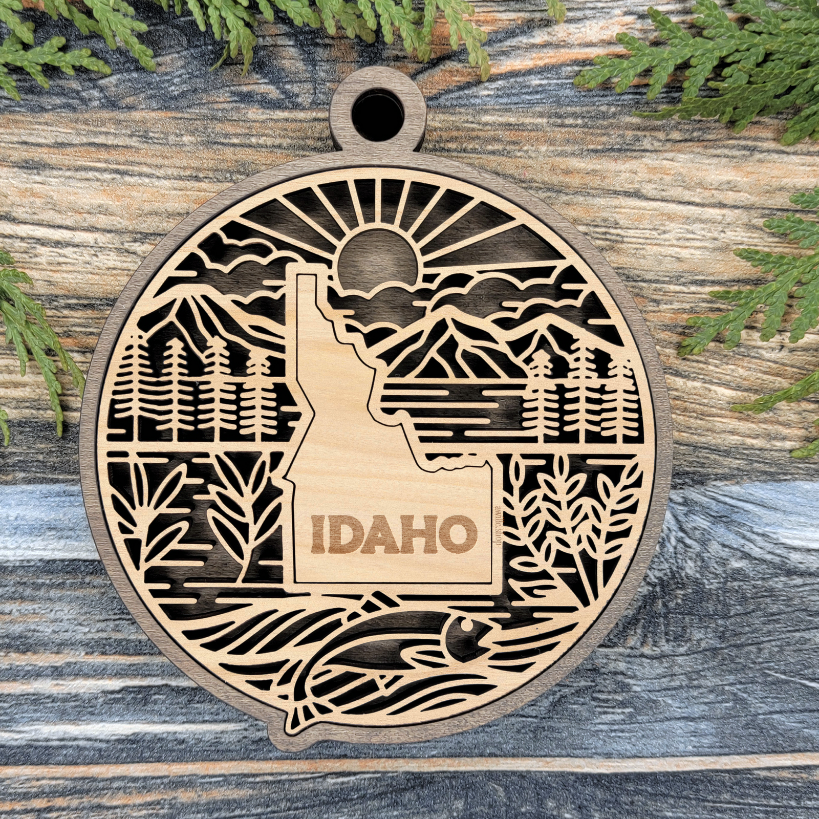 Layered Idaho wood ornament with a laser cut mountains, pine trees, wheat and a trout framing the engraved state shape