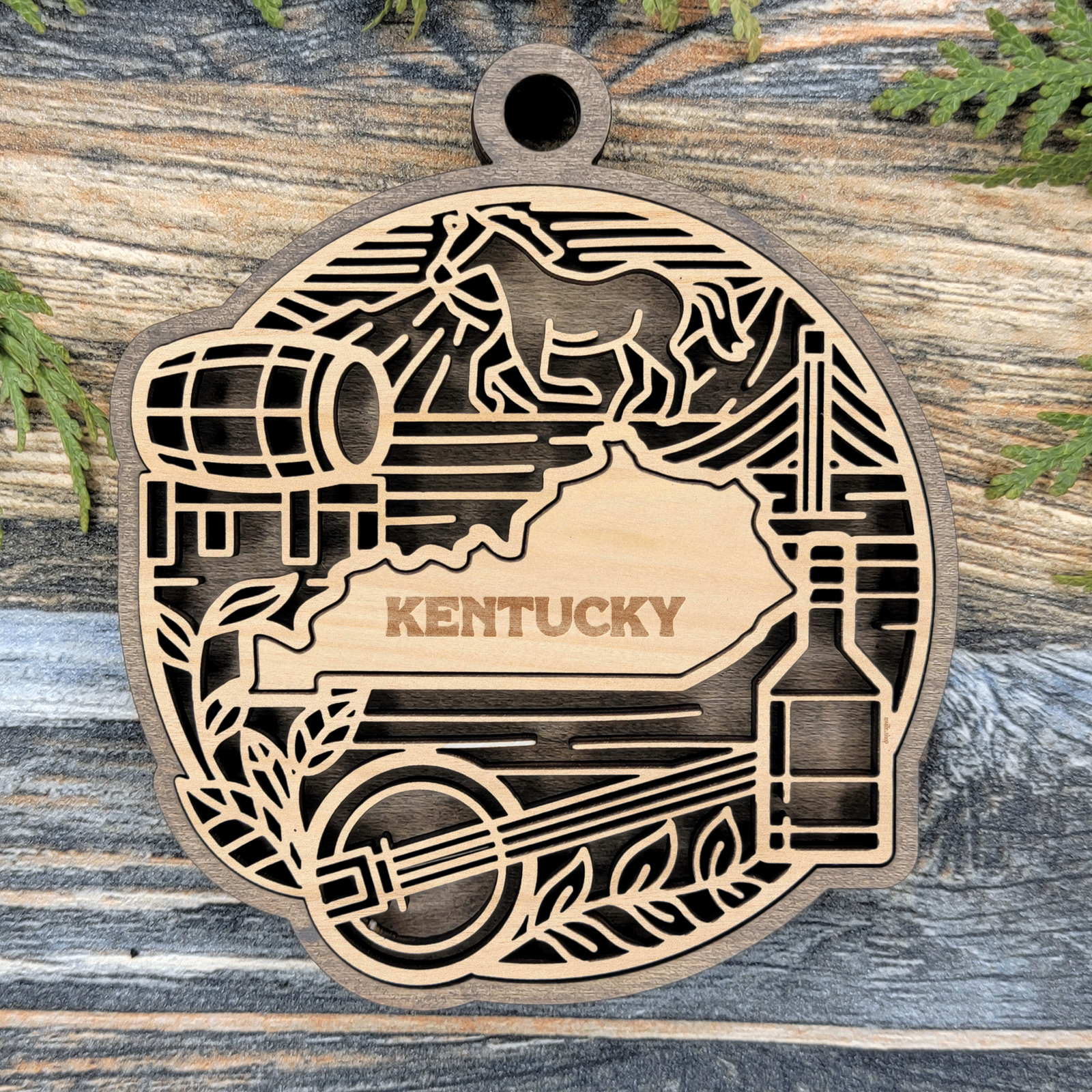 Layered Kentucky wood ornament with a laser cut horse and jockey, a bourbon barrel and bottle, and a banjo framing the engraved state shape