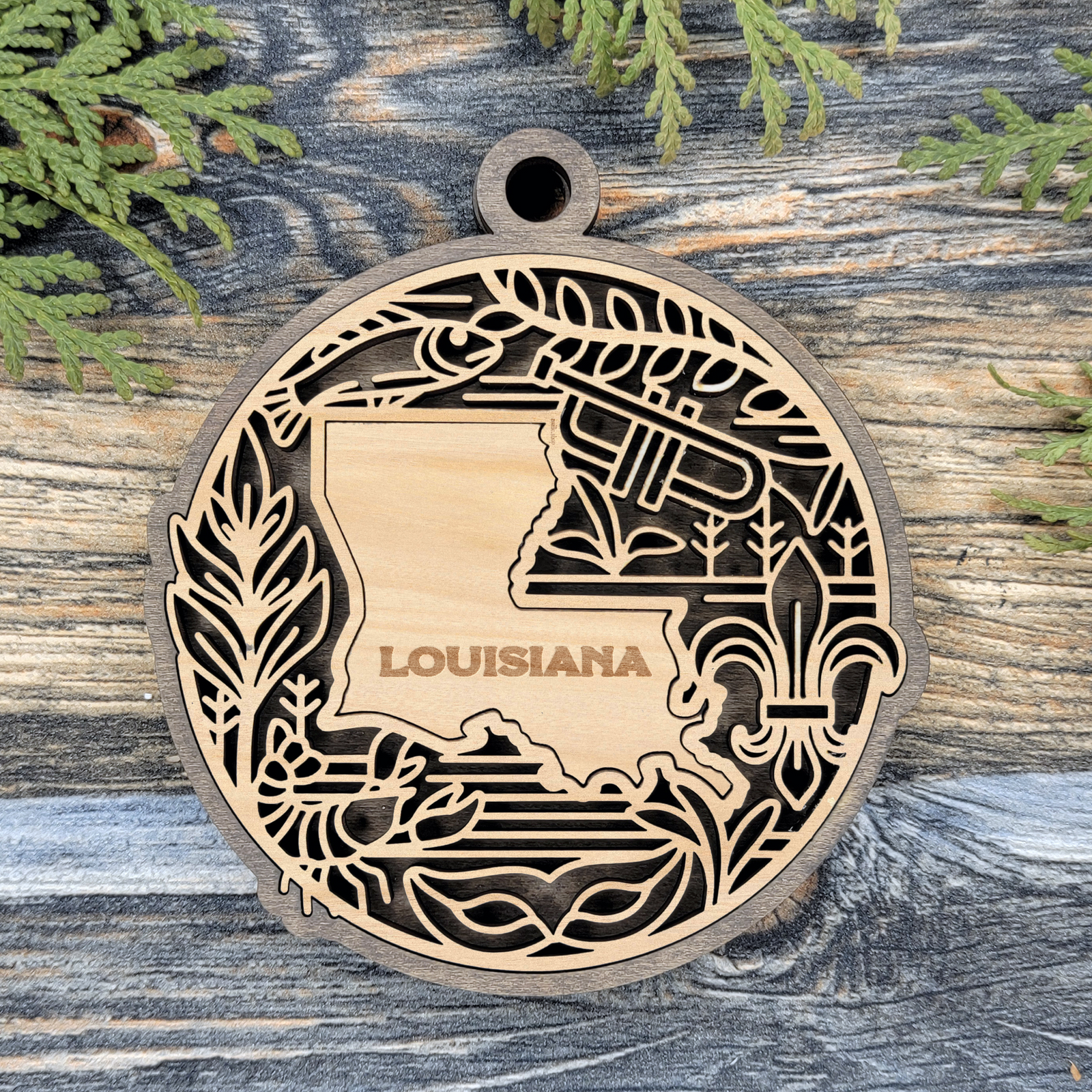 Layered Louisiana wood ornament with a laser cut crawfish, a trumpet, a fleur-de-lis and marsh plants framing the engraved state shape