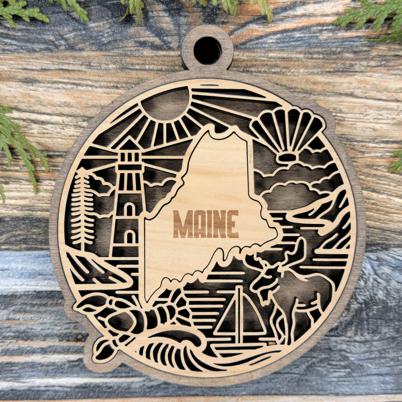 Layered Maine wood ornament with a laser cut lighthouse, a pine tree, a lobster, a sailboat and a moose framing the engraved state shape