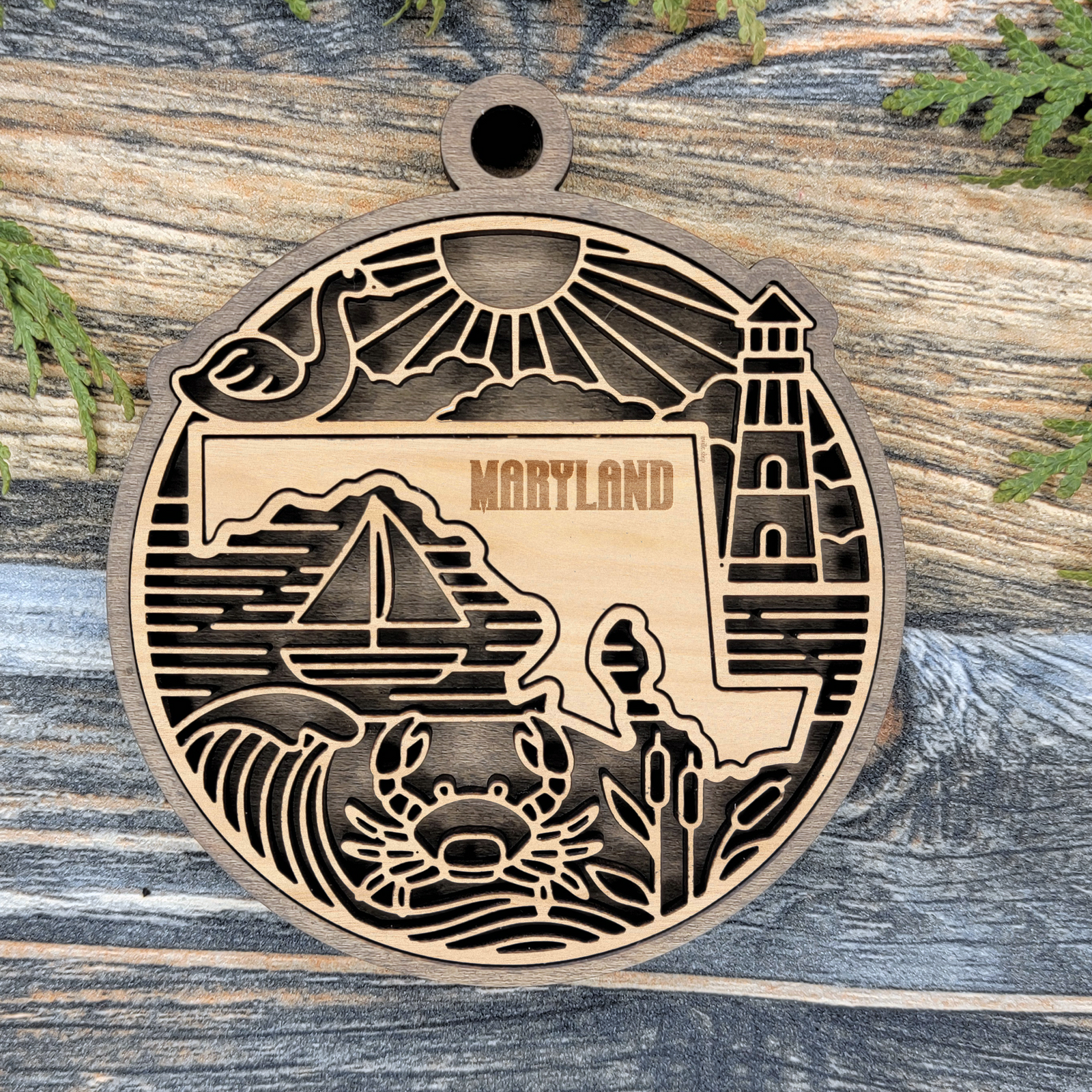 Layered Maryland wood ornament with a laser cut sailboat, a lighthouse, a crab and cattails framing the engraved state shape