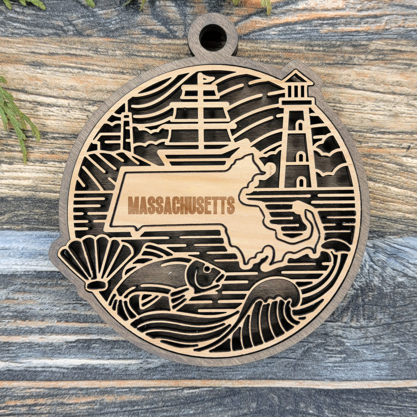 Layered Massachusetts wood ornament with a laser cut sailing ship, a lighthouse, a scallop shell and a fish framing the engraved state shape
