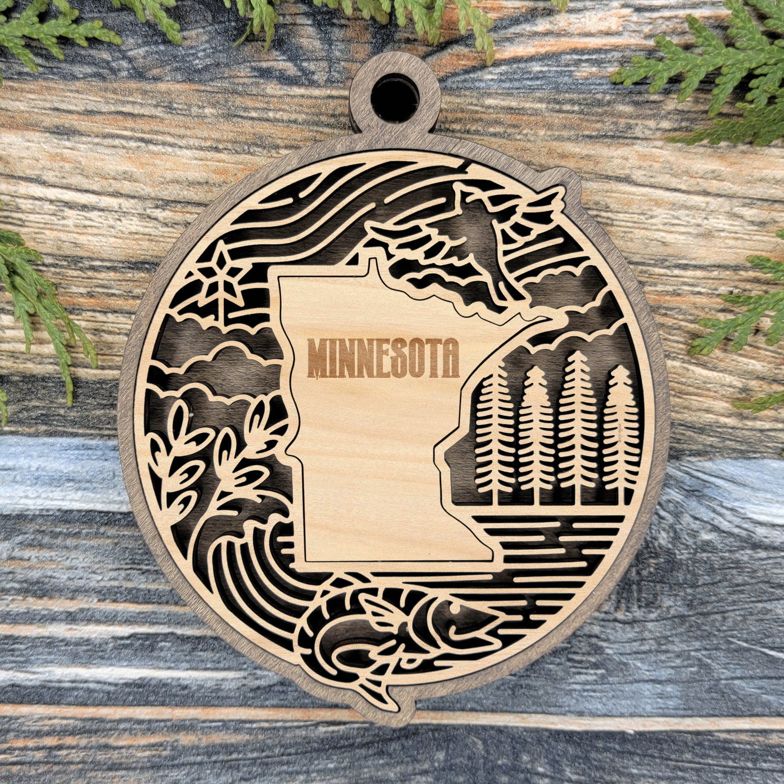 Layered Minnesota wood ornament with a laser cut pine trees, a loon, a fish and a north star framing the engraved state shape