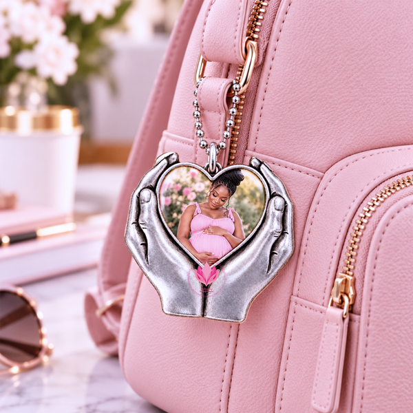 Silver-tone heart-hand photo charm with a portrait, hanging from a pink backpack on a floral dressing table.