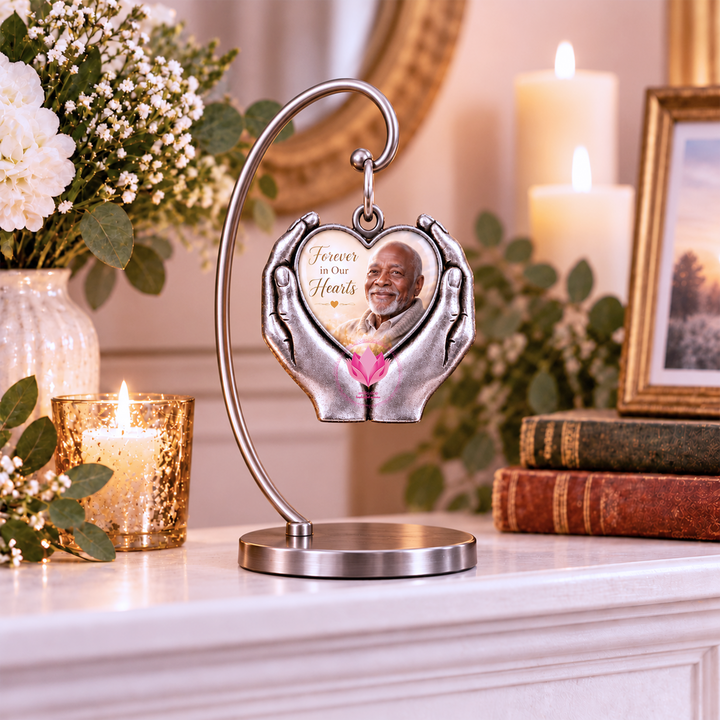 Silver-tone heart-hand photo pendant with a man’s portrait and “Forever in Our Hearts” text, displayed on a stand beside candles.
